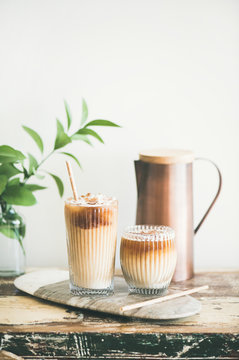 Iced coffee in tall glasses with milk and straws on board over rustic wooden table, white wall, jug and plant branch at background, copy space. Summer refreshing beverage or ice coffee drink concept