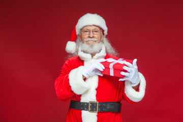 Christmas. Kind Santa Claus in white gloves holds a gift red box with a bow near an ear. He listens. Isolated on red background.