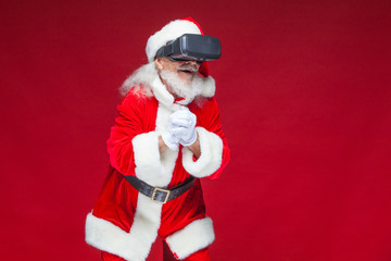 Christmas. Santa Claus in black virtual reality glasses makes gestures with his hands. Surprise, emotion. New technology. Isolated on red background.