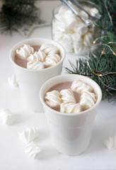 Cocoa with marshmelow in white cups and marshmallows in a glass jar on a white table with spruce branches and garland.