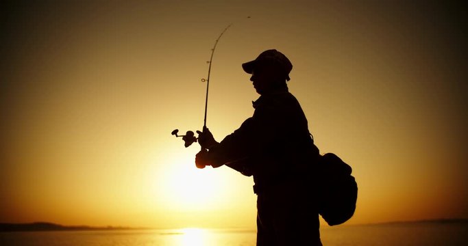 An experienced Fishman catching fish with professional spinning rod from a lake shore. lake in orange sunset 4k