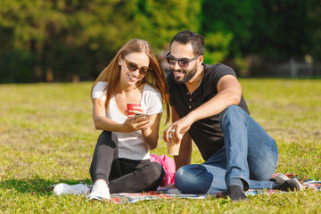 Pretty student-girl in white t-shirt wearing sunglasses and multicultural student-boy in black t-shirt and sunglasses sitting on the grass, drinking coffee and speaking with each other