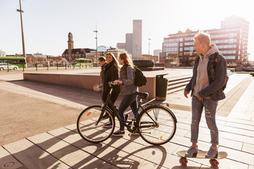 Teenage boy skateboarding while walking with girl holding bicycle on pedestrian zone at city