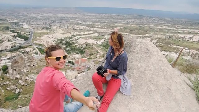 Two young women traveler sits on the edge of the mountain and taking a selfie in the background of the Goreme city, Turkey.