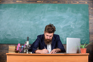 Writing report. Teacher formal jacket sit table classroom chalkboard background. Teachers daily routine paperwork and writing reports. Teacher bearded man writing with pen busy with paperwork