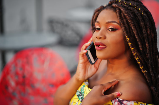 Cute small height african american girl with dreadlocks, wear at coloured yellow dress, sitting at outdoor cafe on red chair and speaking on phone.