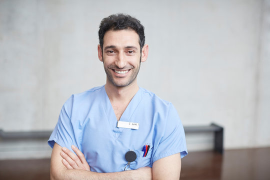 Portrait of smiling young male nurse in blue scrubs standing with arms crossed against wall at hospital