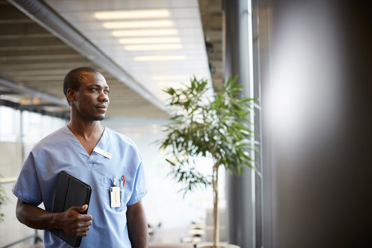 Thoughtful Mid Adult Male Nurse With Digital Tablet Looking Through Window While Standing In Corridor At Hospital