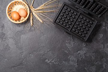 Photo of waffle iron, eggs, wheat spikes on black table