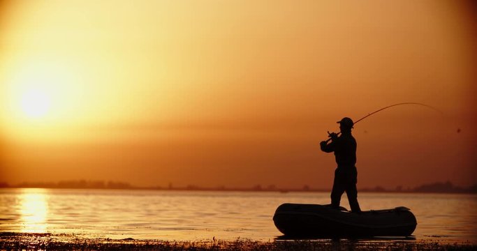 An experienced fisher attempting to catch fish with professional spinnng rod in his rubber boat. Lake in golden sunset 4k