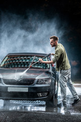 A man with a beard or car washer washes a gray car with a high - pressure washer at night in a shop...