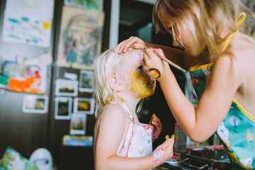 Cute girl painting face of her little sister .