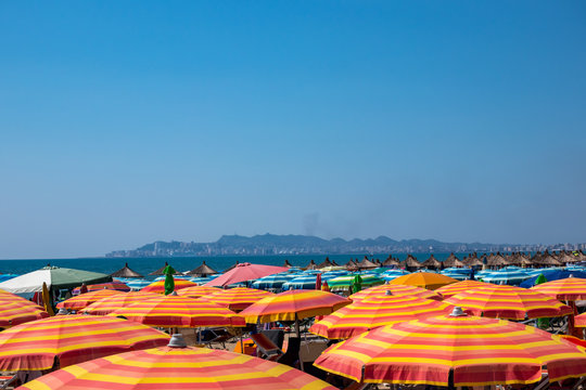 Many colorful sun umbrellas and parasols on a beach in Albania with horizon and city in the background.