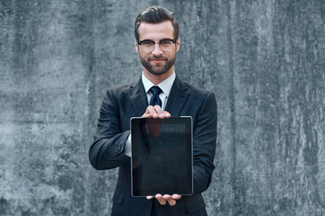 Smiling successful young businessman on a concrete background in a classic suit and tie presents a tablet