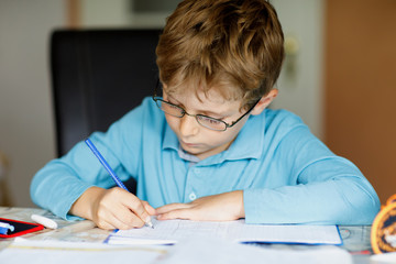 Cute little kid boy with glasses at home making homework, writing letters and doing maths with colorful pens. Little child doing exercise, indoors. Elementary school and education, imagine fantasy