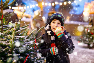 Little cute kid boy eating German sausage and drinking hot children punch on Christmas market....
