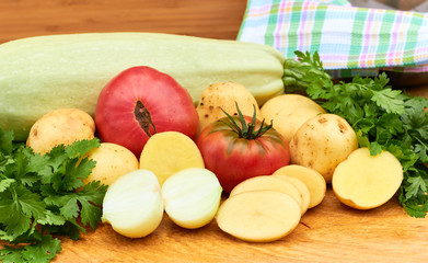 Fresh garden vegetables and herbs: tomatoes, potatoes, onion, parsley, cilantro, zucchini assorted on cutting board on wooden background with kitchen stuff: napkin and bowl, vegan food concept.