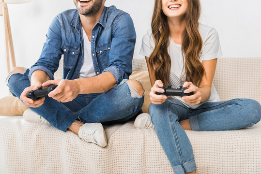 Partial View Of Couple Playing Video Game By Joysticks On Sofa At Home