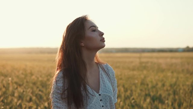 Inspired young woman is enjoying picturesque summer sunset in field. She is standing back to sun and sunlight is putting on her long hair