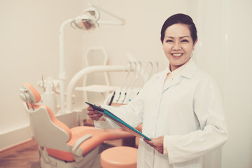 Cheerful Asian adult doctor in dental office holding clipboard and smiling at camera