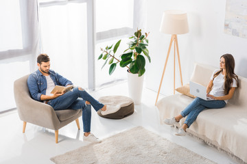 Fototapeta premium high angle view of woman using laptop on sofa while her boyfriend reading book on armchair at home