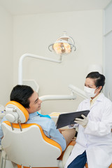 Adult dentist in mask showing tablet to young Asian man in dental chair during visit in office
