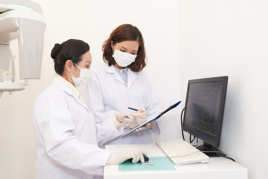Side view of Asian women in white gowns exploring panoramic dental x-ray on computer screen and talking