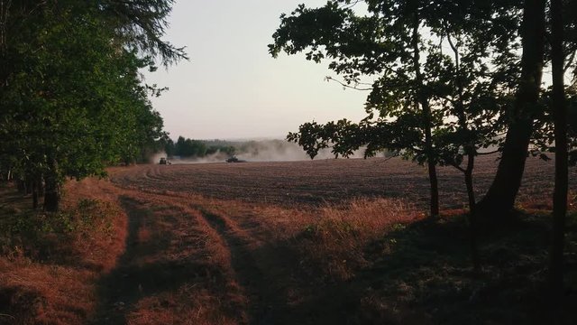 Blick aus dem Wald, zwei Traktoren pfl&uuml;gen Feld und wirbeln Staub auf
