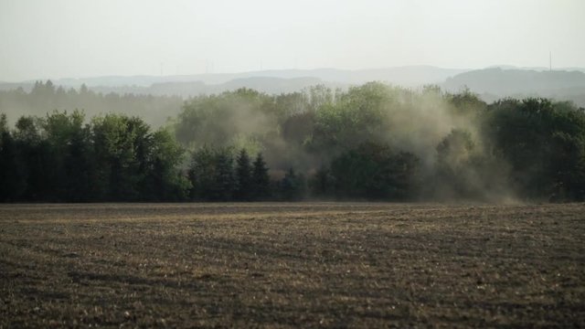 Staubwolken ziehen &uuml;ber abgeerntetes Getreidefeld, dahinter H&uuml;gel mit Mischwald