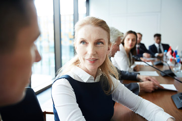 Head and shoulders portrait of mature businesswoman looking at colleague while sitting at meeting table in conference room, copy space