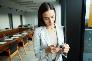 Portrait of elegant businesswoman using smartphone standing by window in office, copy space