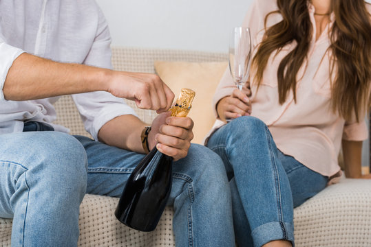 cropped image of man opening champagne bottle while his girlfriend sitting near with glass on sofa at home
