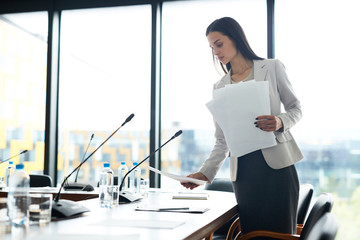 Portrait of elegant businesswoman placing papers on meeting table while prepairing conference room,...