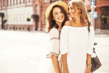 Obraz premium Fashion portrait of two young stylish hippie brunette and blond women models in summer sunny day in white hipster clothes posing on the street background. No makeup