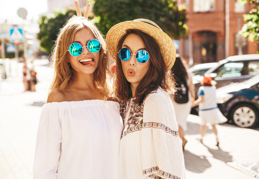Fashion portrait of two young stylish hippie brunette and blond women models without makeup in summer sunny day in white hipster clothes posing on the street background. Going crazy