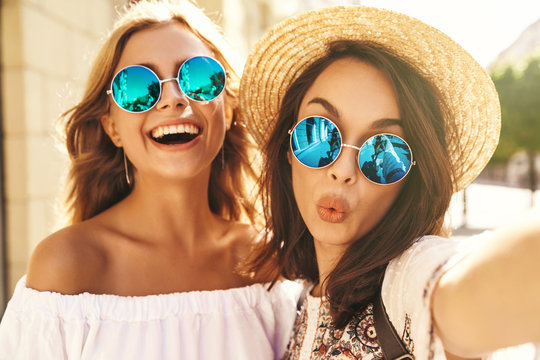 Two young female smiling hippie brunette and blond women models in summer sunny day in white hipster clothes taking selfie photos for social media on smartphone on the street background. Giving kiss