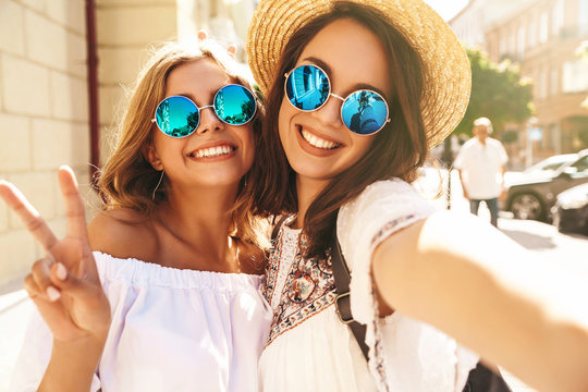 Two young female stylish hippie brunette and blond women models in hipster clothes taking selfie photos for social media on smartphone on the street background