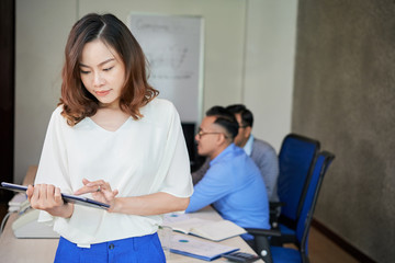 Obraz premium Beautiful Asian woman standing in office room with coworkers on background and using tablet 