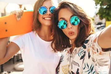 Fotobehang Two young female stylish hippie brunette and blond women models in summer hipster clothes taking selfie photos for social media on smartphone on the street background. With colorful penny skateboards © halayalex