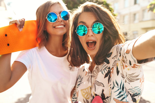 Two female stylish hippie brunette and blond women models in hipster clothes taking selfie photos for social media on smartphone on the street background