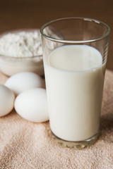 glass of milk, white chicken eggs and bowl with flour on brown tablecloth