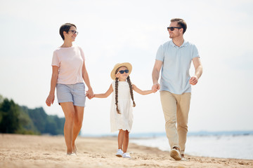 Full length portrait of  happy family holding hands with cute little girl while enjoying walk on beach during Summer vacation