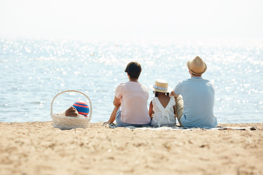 Back View Portrait Of Blissful Modern Family Sitting On Beach And Looking At Sea Enjoying Picnic During Summer Vacation, Copy Space