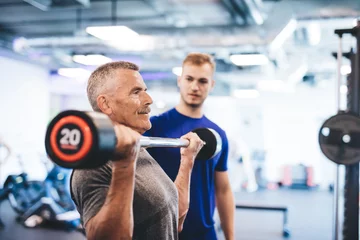 Fototapeten Older man lifting weights, supervised by gym assistant. © Photocreo Bednarek
