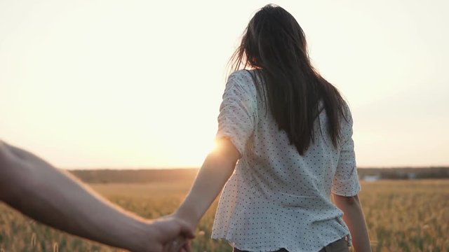 Young charming girl is holding hand of her boyfriend, stepping on field in sunset in summer. She is looking to him and turning head, back view