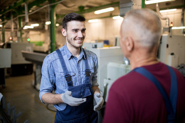 Happy young worker in overalls and gloves speaking to his mature experienced colleague in workshop