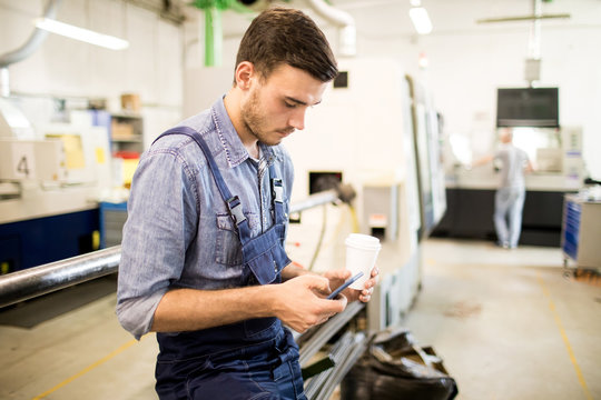 Young engineer with mobile phone and glass of coffee texting by industrial machine in workshop