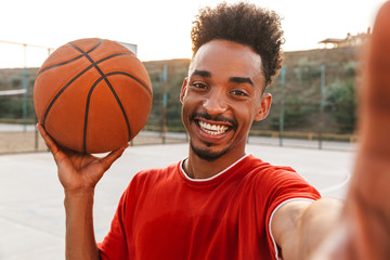 Portrait of smiling african american man holding ball and taking selfie, while playing basketball...