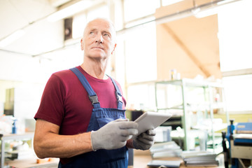 Mature professional in workwear and gloves browsing in the net during working day at factory