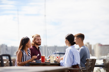 Two young couples in casualwear sitting by table in outdoor cafe and discussing news by lunch